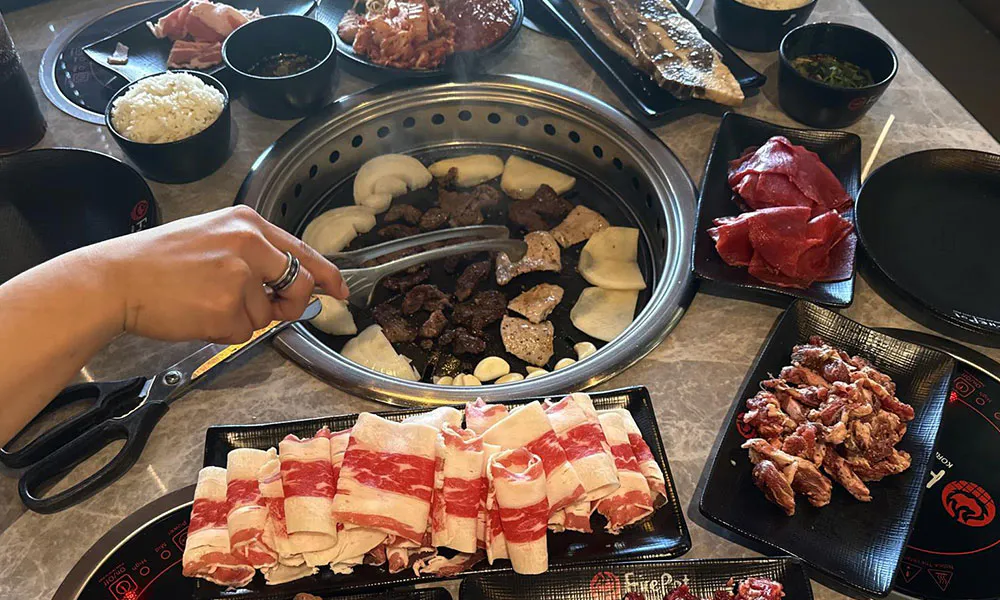 Korean BBQ table setup with grill, assorted raw meats, rice bowl and hand using tongs at FirePot Korean BBQ & Hot Pot, an Asian Restaurant in Grand Forks